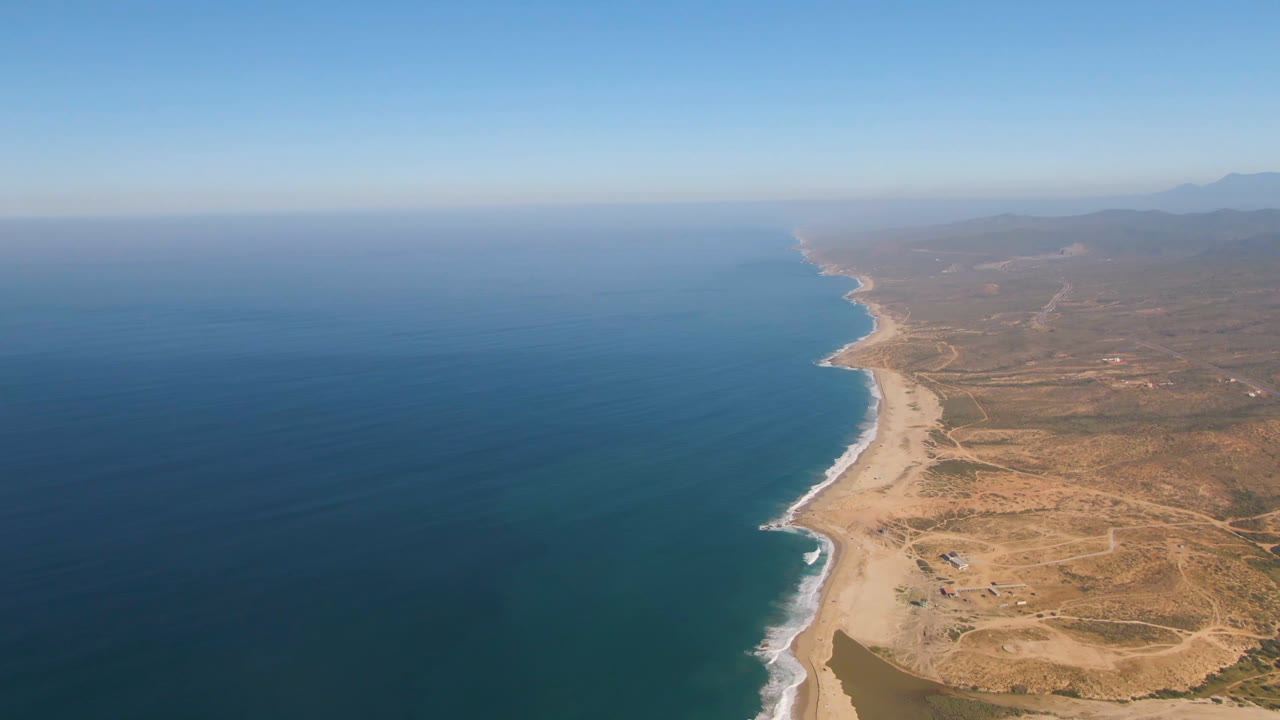 High drone view over Migrino, Baja California Sur with blue ocean to the left, pale beach curving along dry coastal terrain, distant hills, and no visible people.