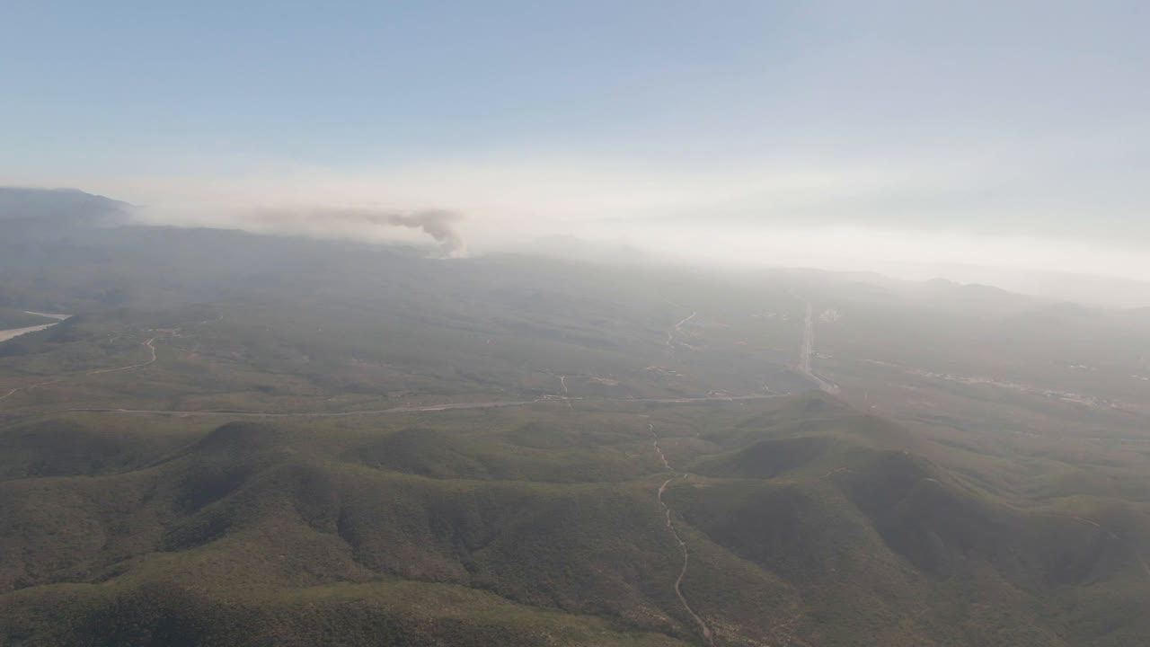 Wide aerial landscape in Migrino, Baja California Sur, showing rounded hills in the foreground, faint road lines through the valley, distant smoke, and no visible people.