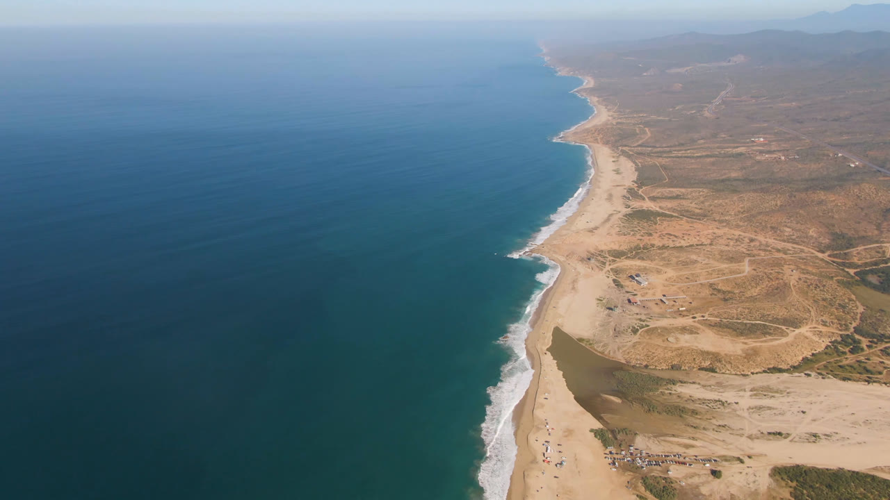 High aerial view over Migrino, Baja California Sur, with deep blue ocean, a pale sand beach, a small inlet near shore, dry scrubland inland, distant hills, and no visible people.