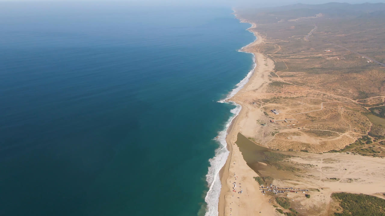 Aerial coastline frame from Migrino showing blue water, a narrow white surf edge, light sand, inland desert tracks, a small lagoon shape near the beach, and no visible people.