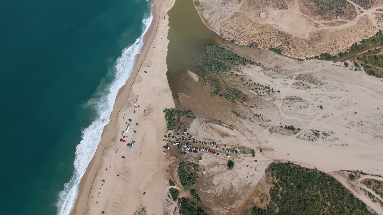 Overhead beach, surf, lagoon water, dunes, and parked vehicles