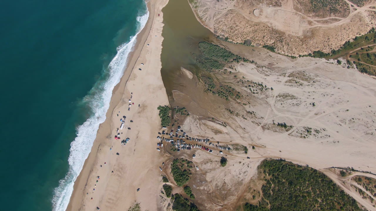 Aerial frame over Migrino beach with the surf line slightly farther down the shore, open sand in the center, lagoon water near the top, parked vehicles to the right, and no clearly visible people.