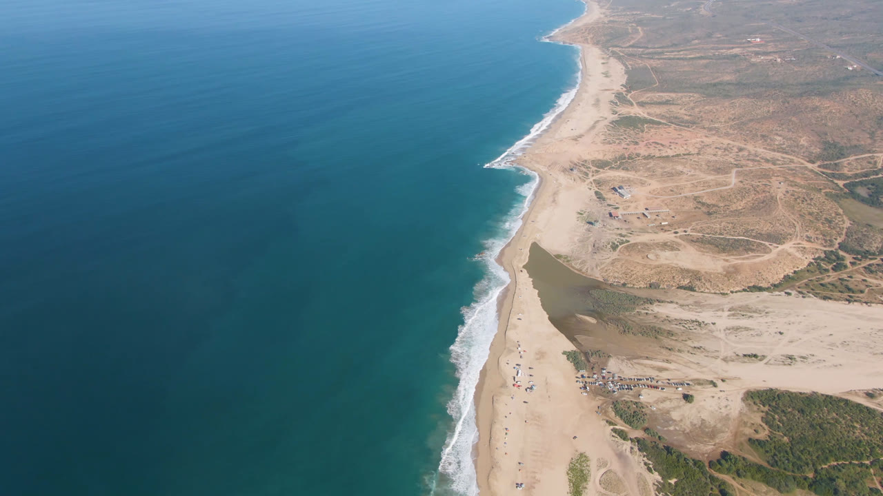 High aerial view of blue ocean, sandy beach, surf, and dry coastal terrain.