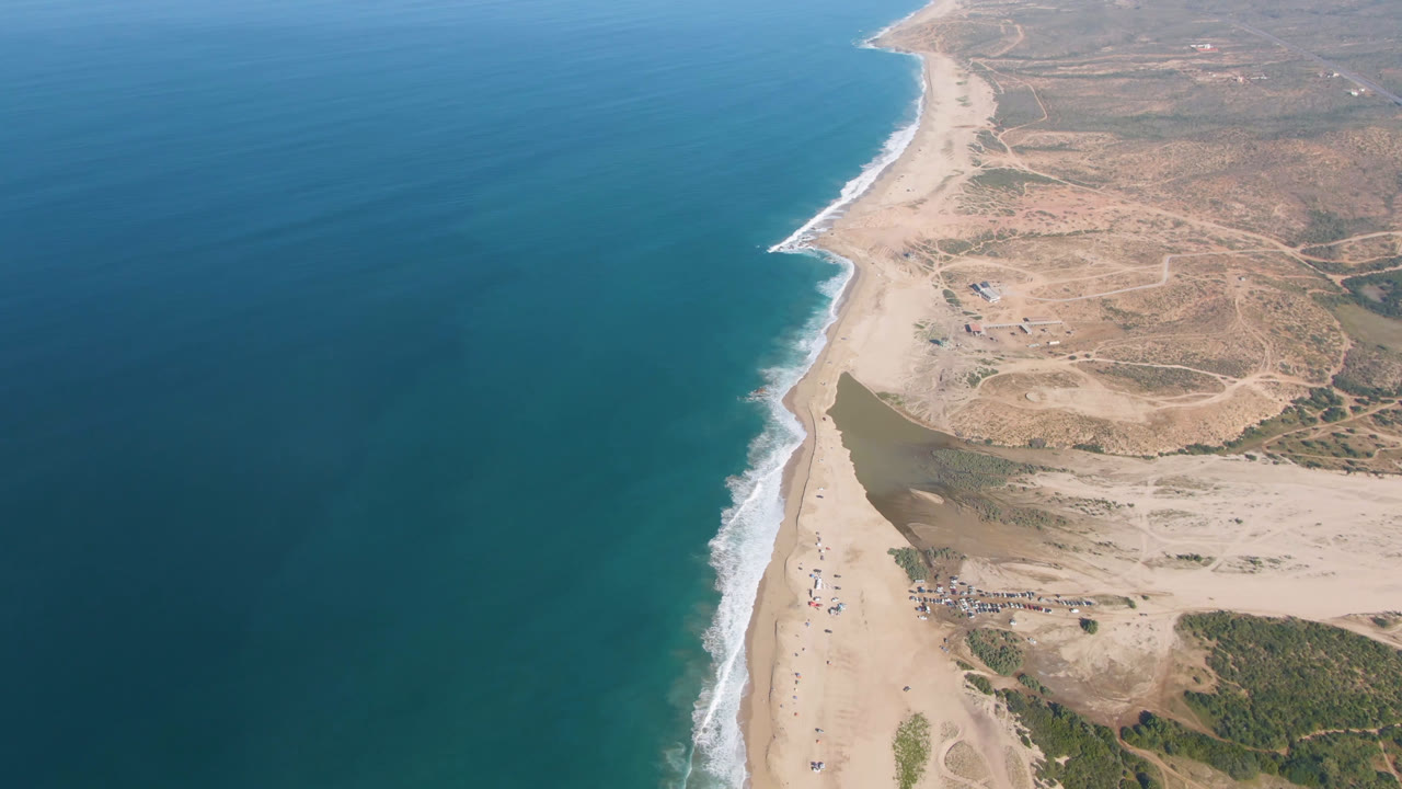 Aerial view of the Migrino shoreline with deep blue water, a diagonal strip of sand, breaking waves, inland dirt tracks, and small objects near the beach access area.