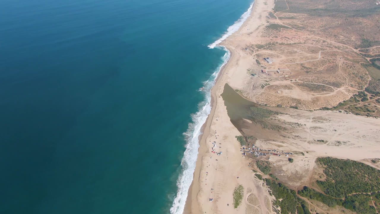 Aerial view of blue ocean, sandy shoreline, surf, and dry coastal terrain.