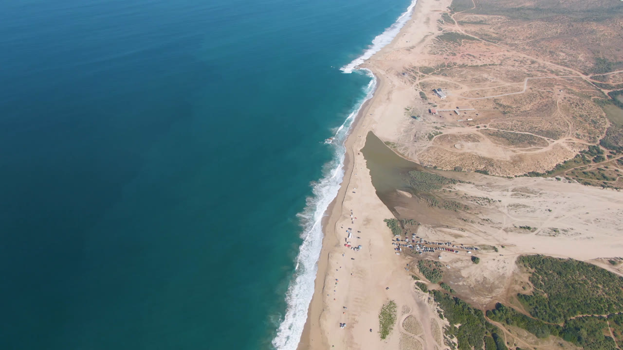 Drone frame over Migrino showing the surf line running diagonally, open blue water, pale beach sand, arid inland tracks, and no visible people.