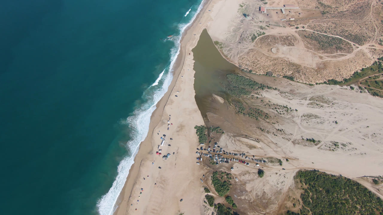 Aerial frame over Migrino, Baja California Sur, Mexico, showing teal ocean, white surf, pale beach sand, a dark coastal body of water, scrubland, parked vehicles, and no visible people.