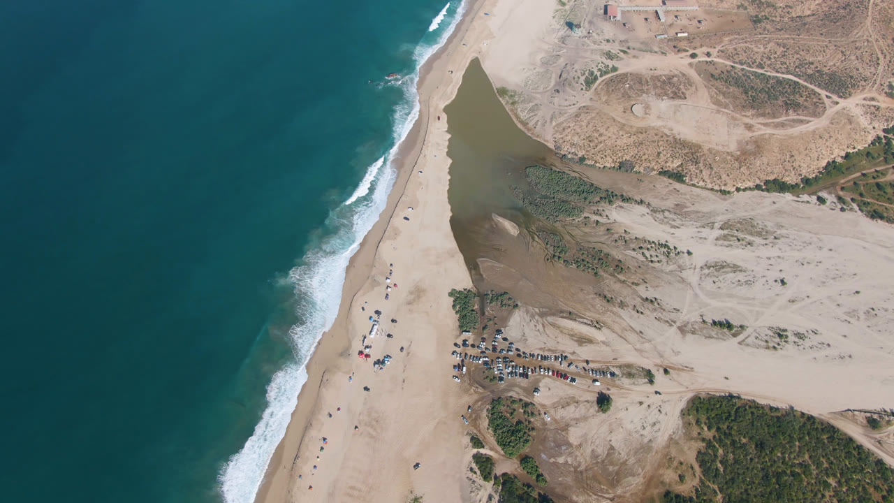 Second aerial frame over Migrino with the surf line slightly lower in view, the dark water inlet beside the beach, dry open sand, clustered parked vehicles, and no visible people.