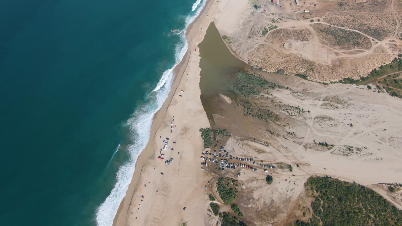 Drone view of beach shoreline, inland water, sand flats, and vehicle cluster.