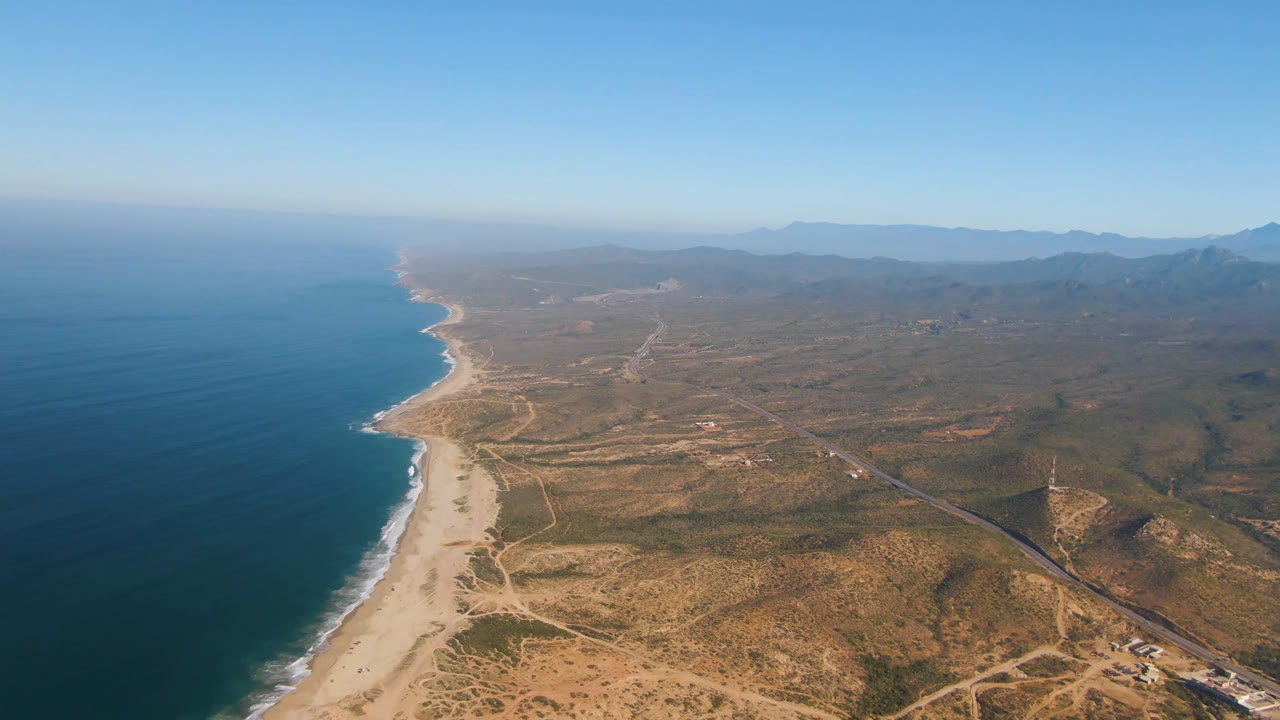 Wide aerial frame over Migrino, Baja California Sur, with blue ocean at left, a long pale beach below, scrubland and roads inland, distant mountains, and no visible people.