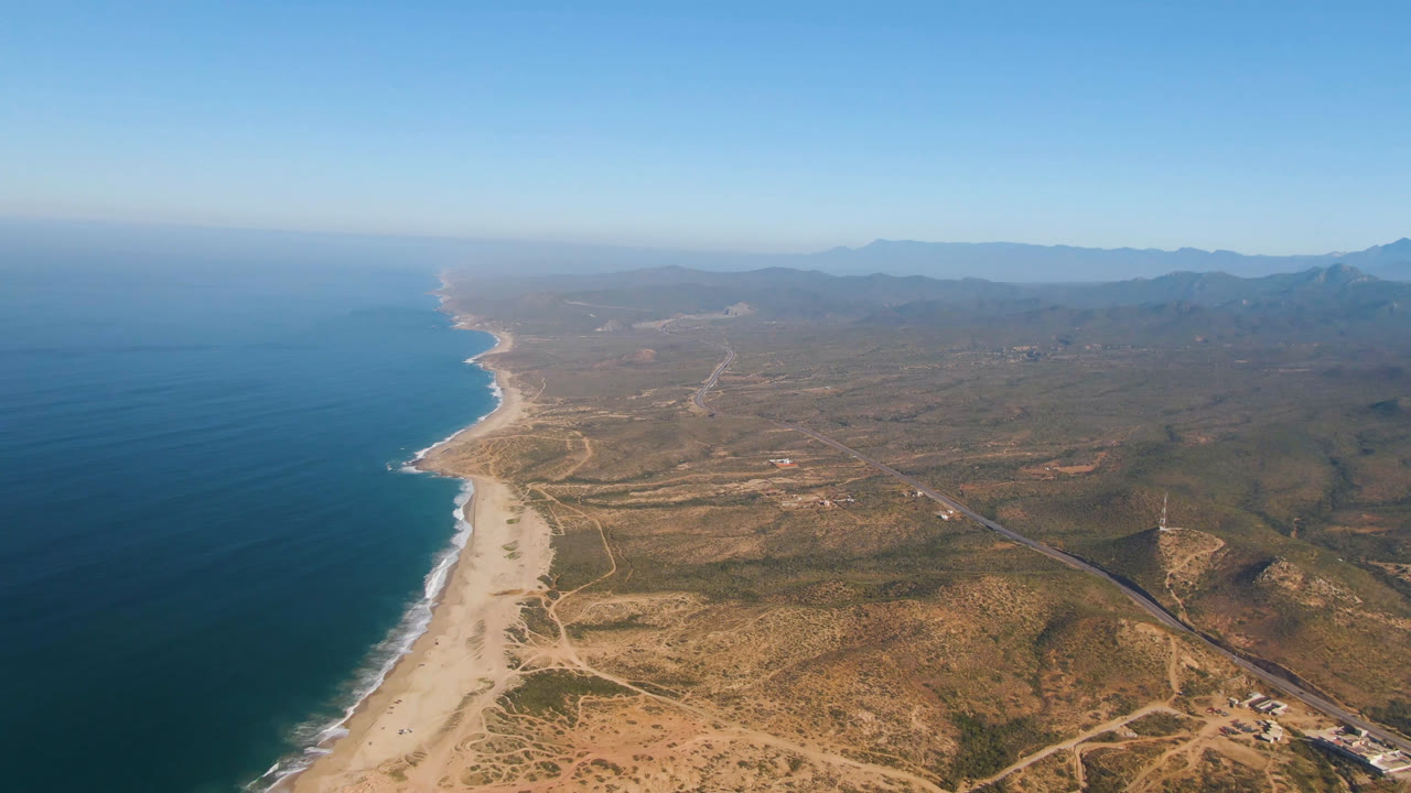 High aerial of a long beach beside blue water and open scrubland.