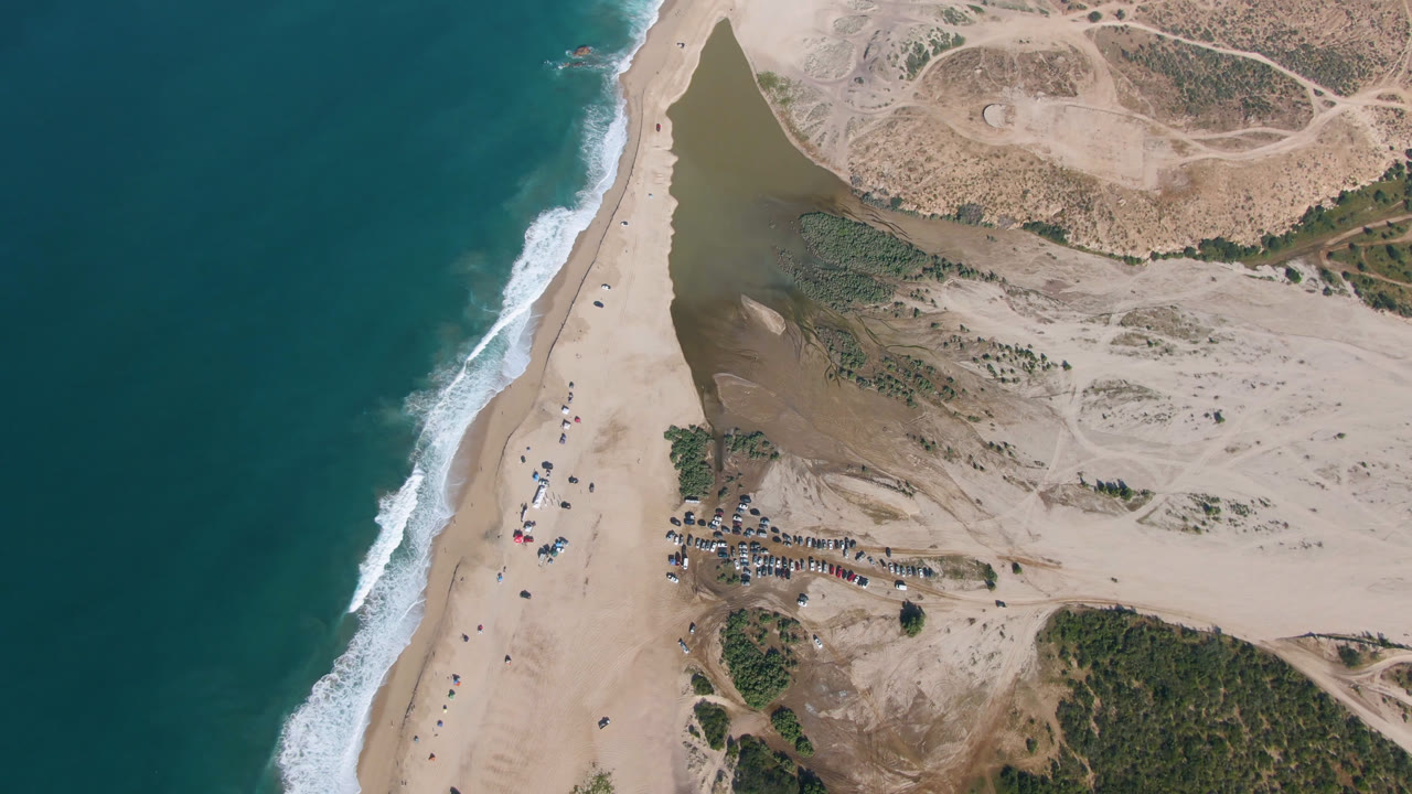 High aerial view of beach, ocean, dunes, lagoon water, vehicles, and small distant people.