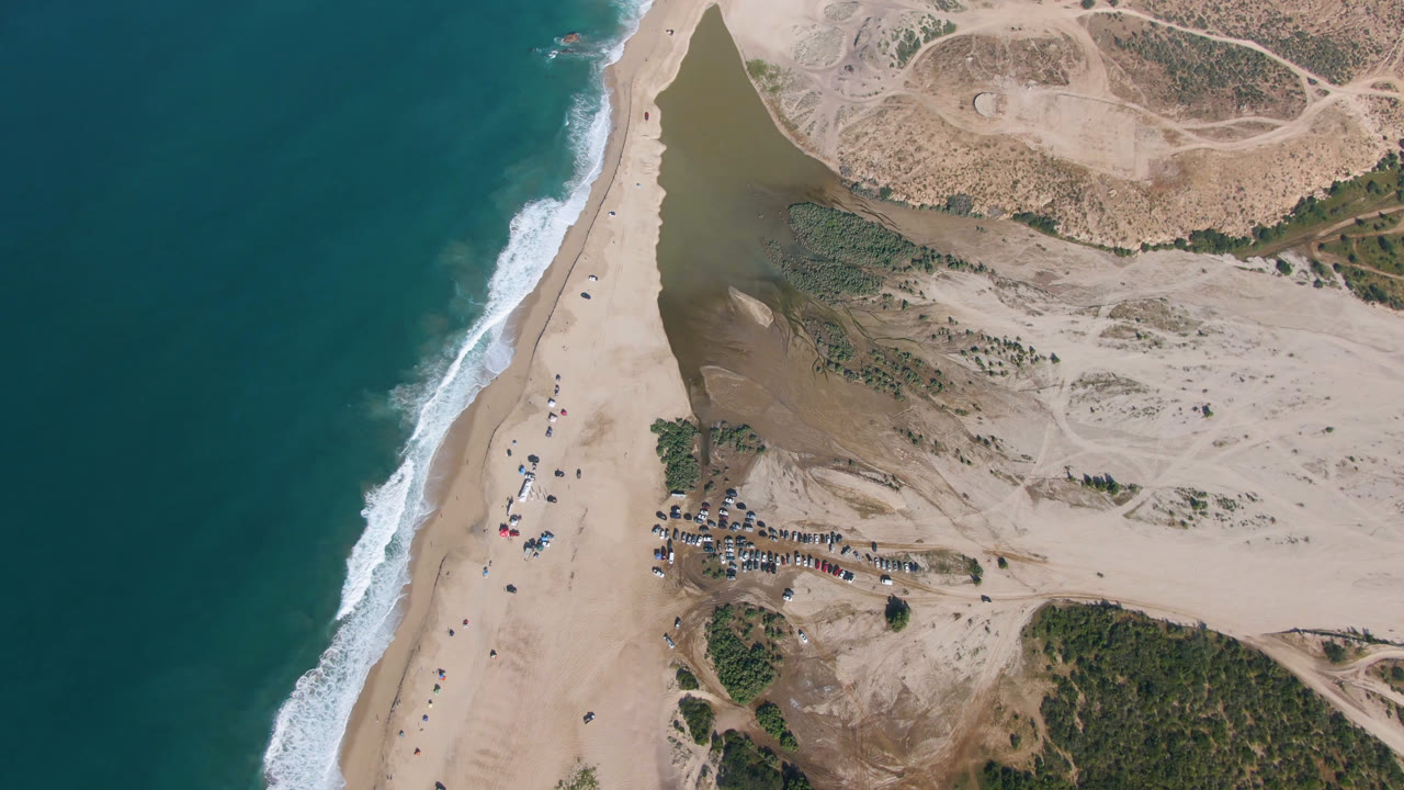 Second aerial frame over Migrino with the surf line pressed along the left beach edge, small distant people on the sand, vehicles grouped near the lower center, and dry tracks across the dunes.