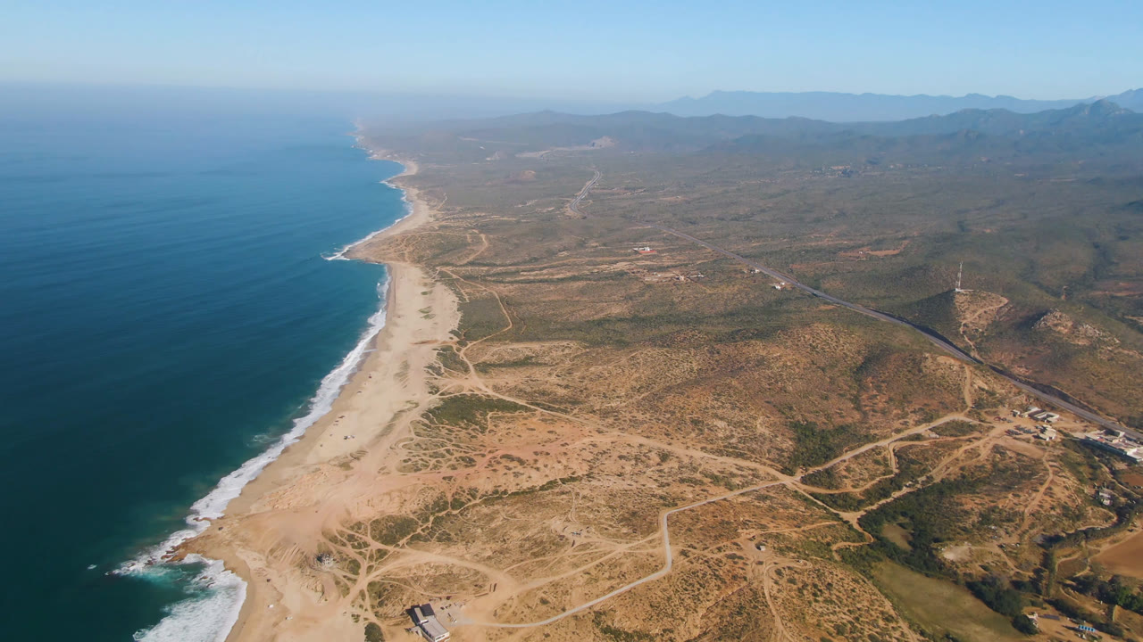 Wide aerial view over Migrino, Baja California Sur, with dark turquoise ocean on the left, surf along a long sandy beach, dry inland tracks, distant hills, and no visible people.