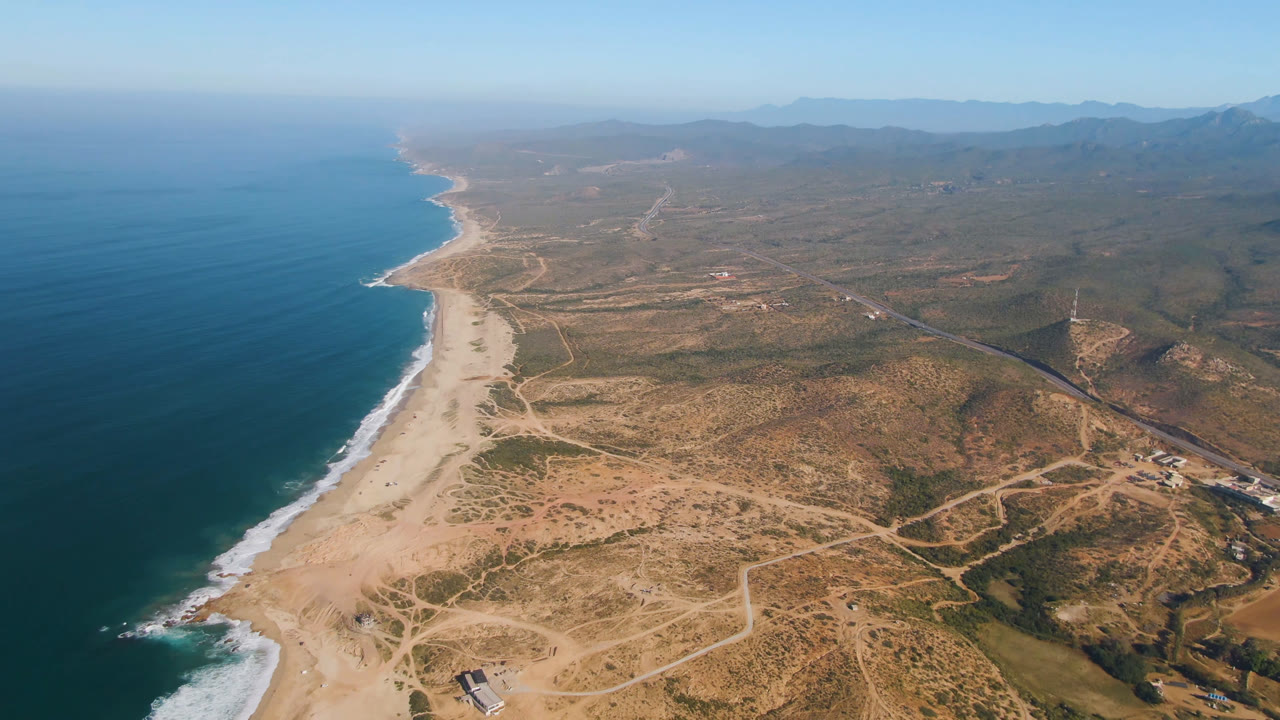 Aerial frame of Migrino coastline with the beach curving into the distance, ocean filling the left side, sandy access roads across the dry terrain, faint buildings, and no visible people.
