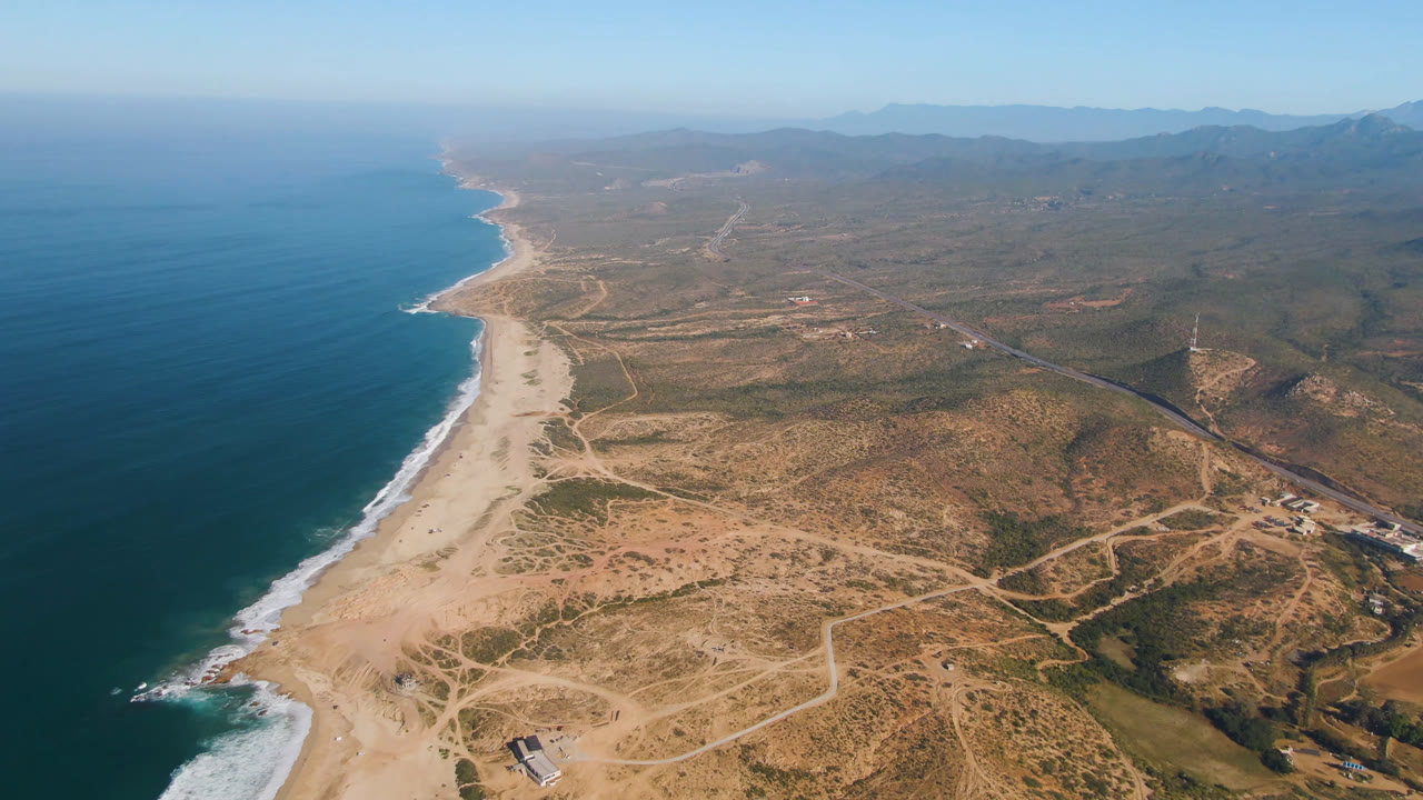 High aerial of ocean shoreline, broad sand, desert tracks, scattered structures, and mountains.