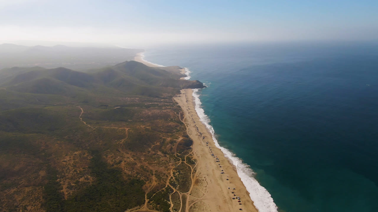 Opening frame over Migrino, Baja California Sur, showing a diagonal sand beach beside blue ocean, scrub-covered land, pale surf lines, distant mountains, and no visible people.