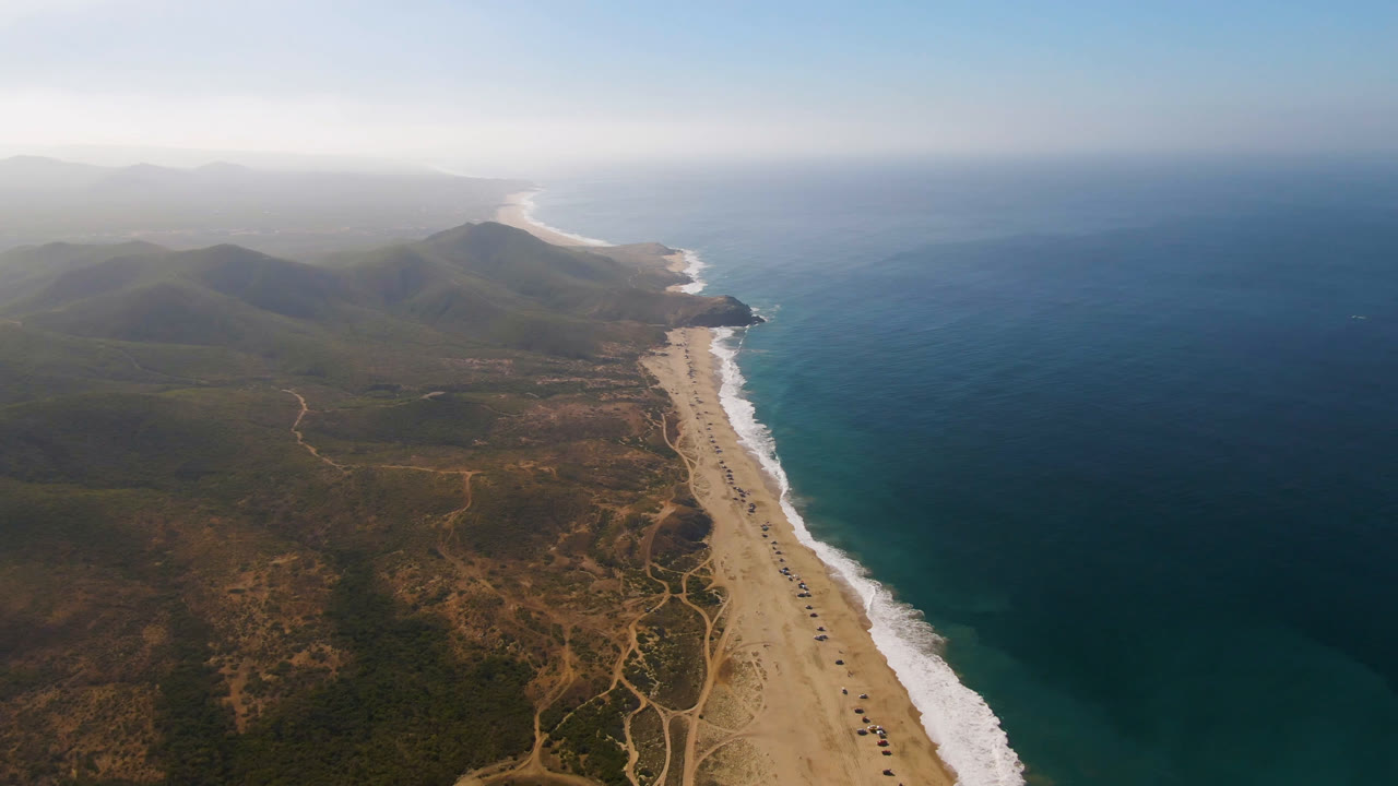 Second frame over Migrino, Baja California Sur, with the ocean occupying the right side, the beach and surf running diagonally, dry tracks inland, hazy mountain ridges, and no visible people.