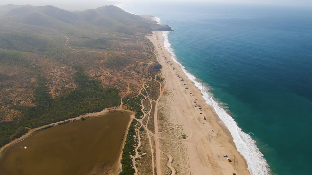Aerial view of sandy coastline, lagoon, surf, scrubland, and mountains.