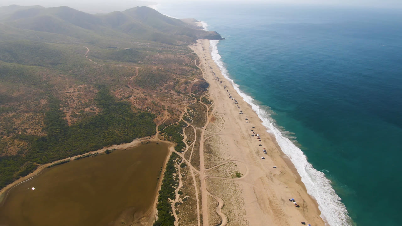 Aerial view over Migrino showing the shoreline running diagonally through frame, blue water to the right, inland tracks and lagoon to the left, and no visible people.