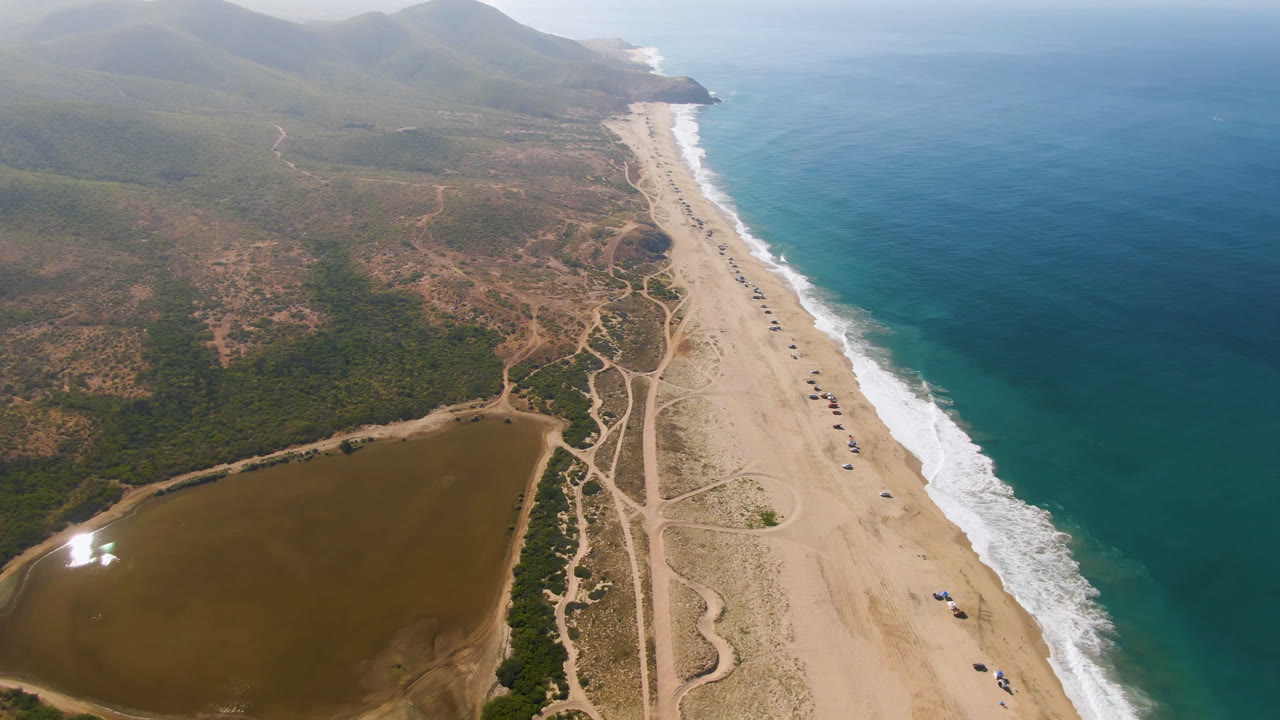 Aerial view over Migrino, Baja California Sur, Mexico, with a brown inland lagoon at lower left, a long sandy beach, turquoise ocean surf at right, hazy hills in the distance, and no visible people.
