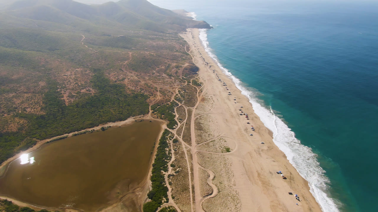 High drone-style frame over Migrino showing the lagoon slightly farther left, dirt roads running along the sand, small parked vehicles near the shoreline, ocean on the right, and no visible people.
