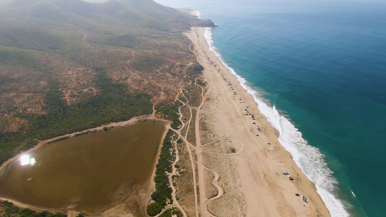 Aerial coastline with bright lagoon reflections, beach tracks, ocean water, surf, and hills.