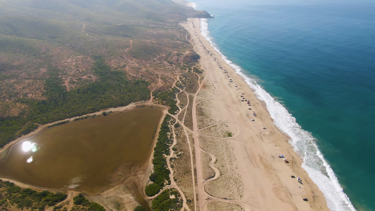 Aerial view over Migrino, Baja California Sur, Mexico, showing a sandy beach between blue ocean surf and a brown inland body of water, with no visible people.