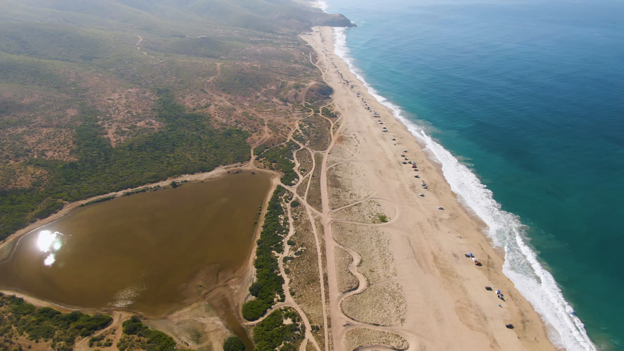 High drone frame over Migrino coastline with pale sand, winding dirt tracks, green scrubland, ocean surf to the right, and no visible people.