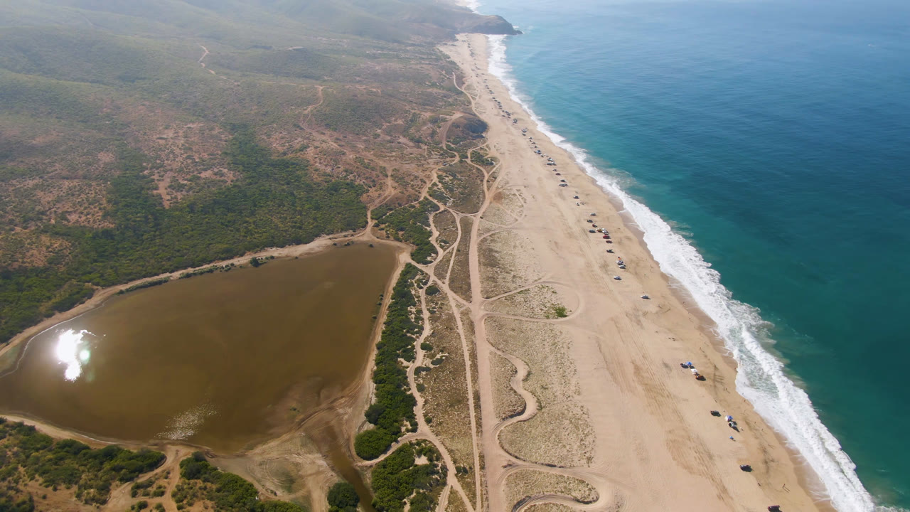Drone view of empty shoreline beside scrubland and reflective inland water.