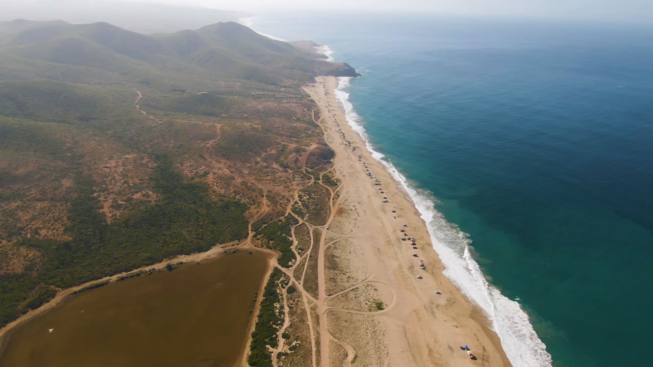 Aerial frame in Migrino showing the coastline running toward hazy mountains, with white surf on the right, sandy beach through the center, scrubland access roads on the left, and no visible people.