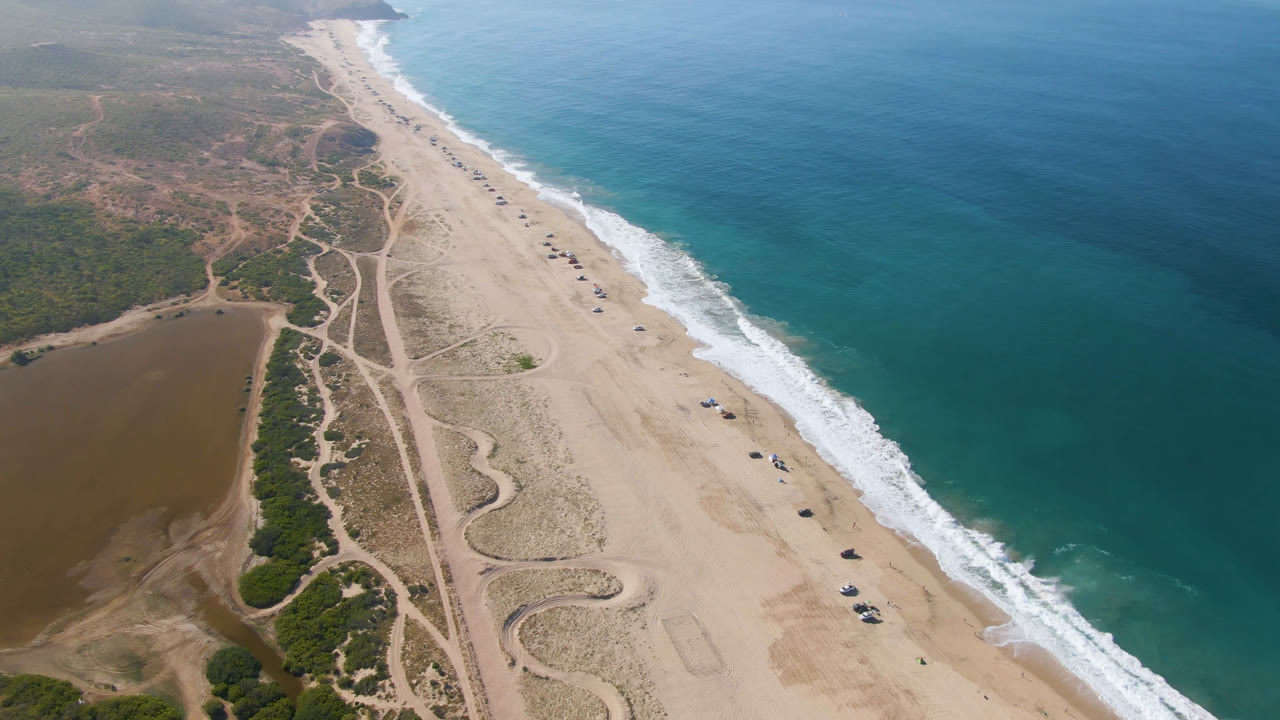 High aerial view over Migrino, Baja California Sur, with a long sandy beach, blue-green ocean, white surf, dune roads, a brown lagoon, and no visible people.