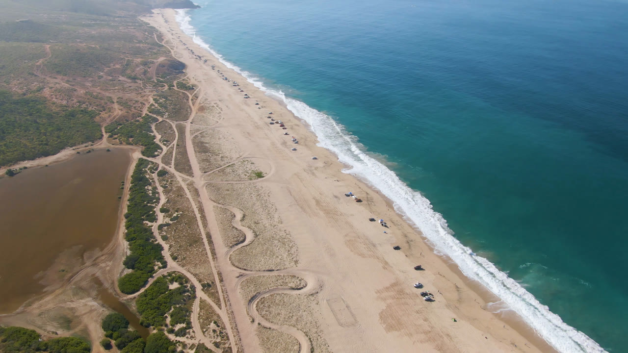 Drone frame over Migrino showing the surf line running along a broad sand beach, with inland dirt tracks, scrub vegetation, small beach vehicles, and no visible people.