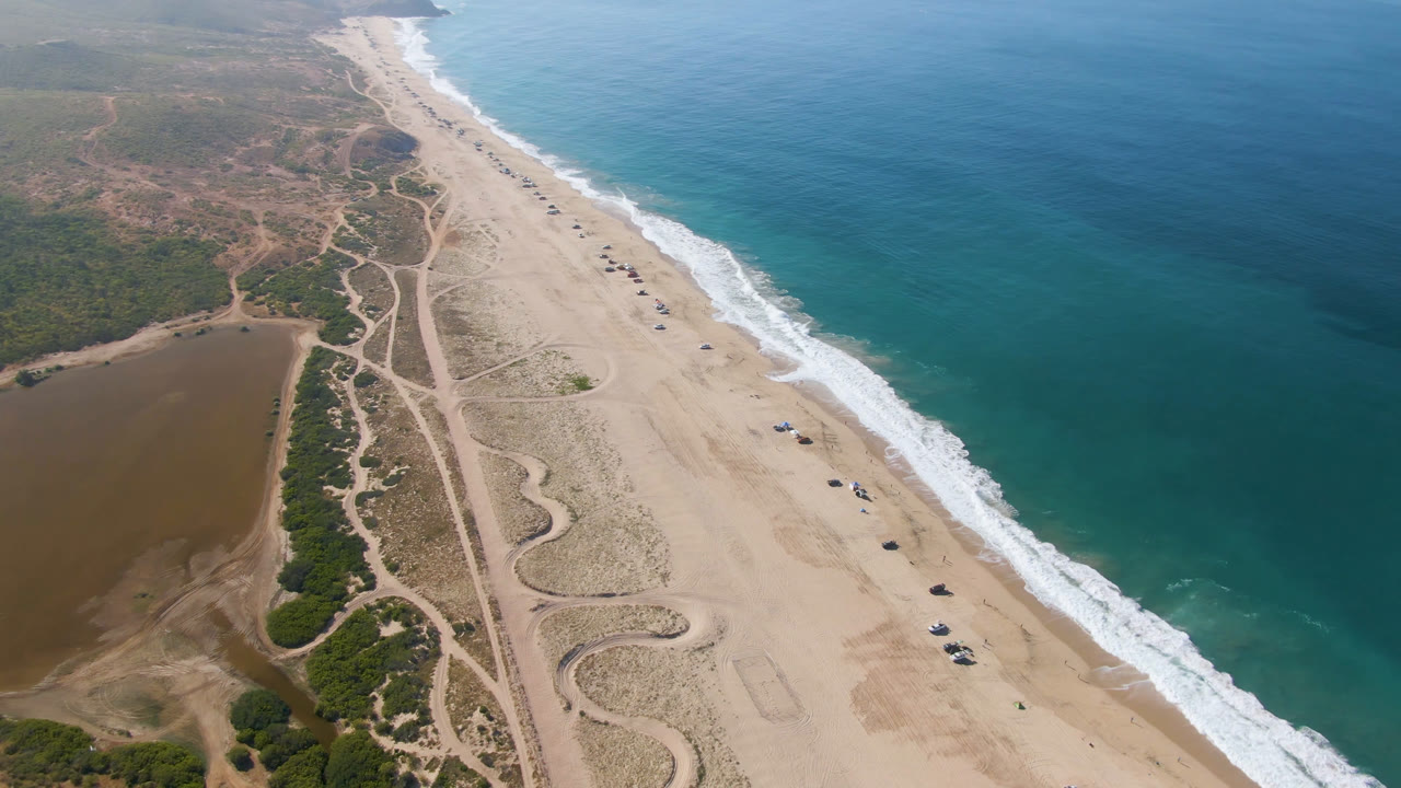 Aerial coastline view with turquoise water, white waves, sand tracks, and parked vehicles.