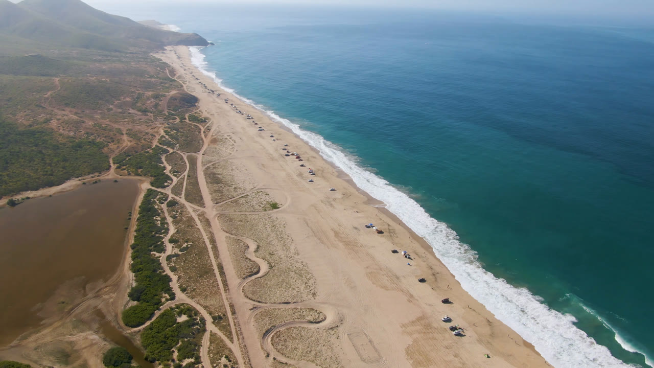 Opening aerial frame over Migrino, Baja California Sur, with a long sandy beach, white surf, blue ocean, dirt tracks, a brown water area inland, distant hills, and no visible people.