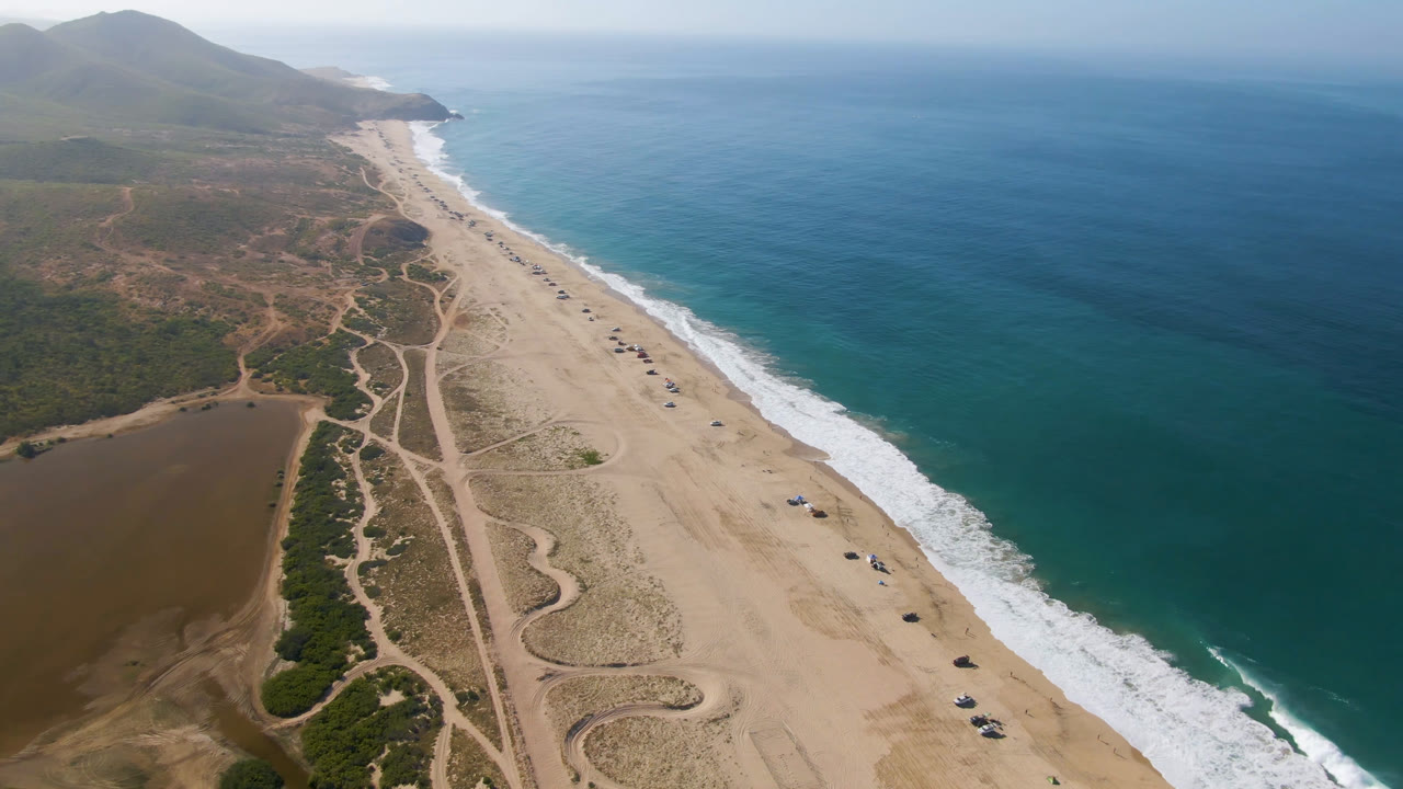 Aerial frame over Migrino beach showing the surf line closer to center, a diagonal strip of sand, winding dirt access tracks, sparse vehicles on the beach, coastal hills, and no visible people.