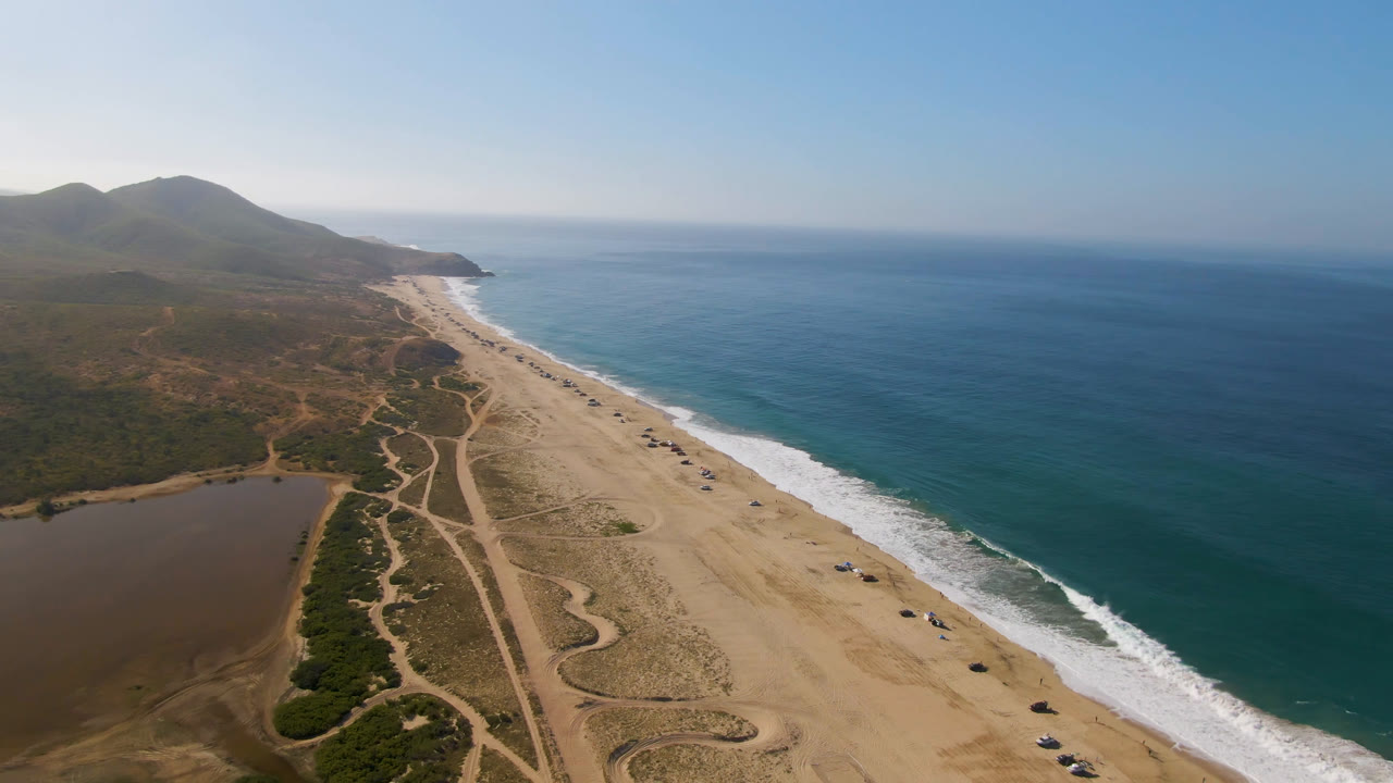 Early aerial frame over Migrino, Baja California Sur, showing a long sand beach beside blue ocean, a brown lagoon at lower left, inland dirt tracks, distant mountains, and no visible people.