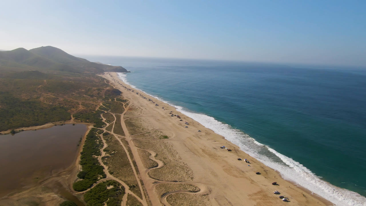 Aerial view over Migrino coastline with the ocean filling the right side, pale beach through the center, lagoon and brush at lower left, mountains on the horizon, and no visible people.