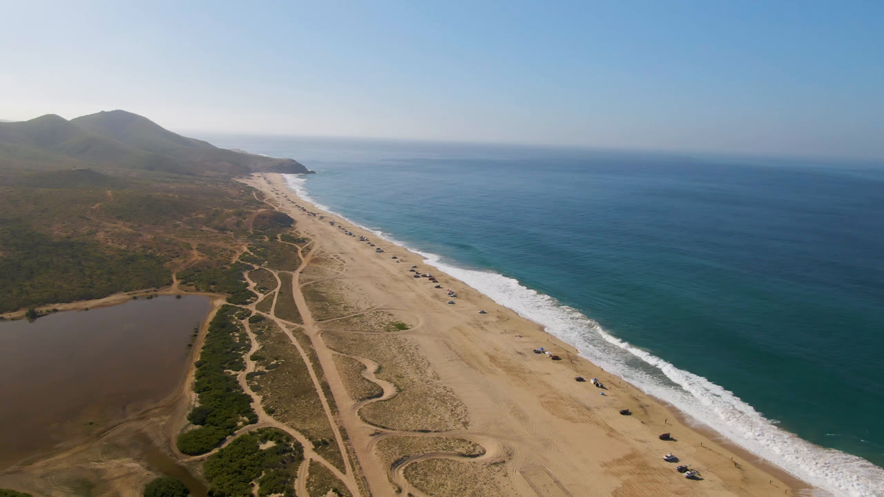 Mid-clip aerial frame from Migrino, Baja California Sur, with white surf bands along the sand, inland brush and tracks, a lagoon edge in the lower left, distant hills, and no visible people.