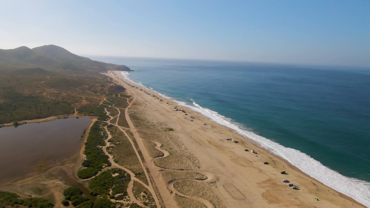 A high aerial frame over Migrino, Baja California Sur shows the beach between blue ocean and a brown inland body of water, with dirt tracks through scrub and no visible people.