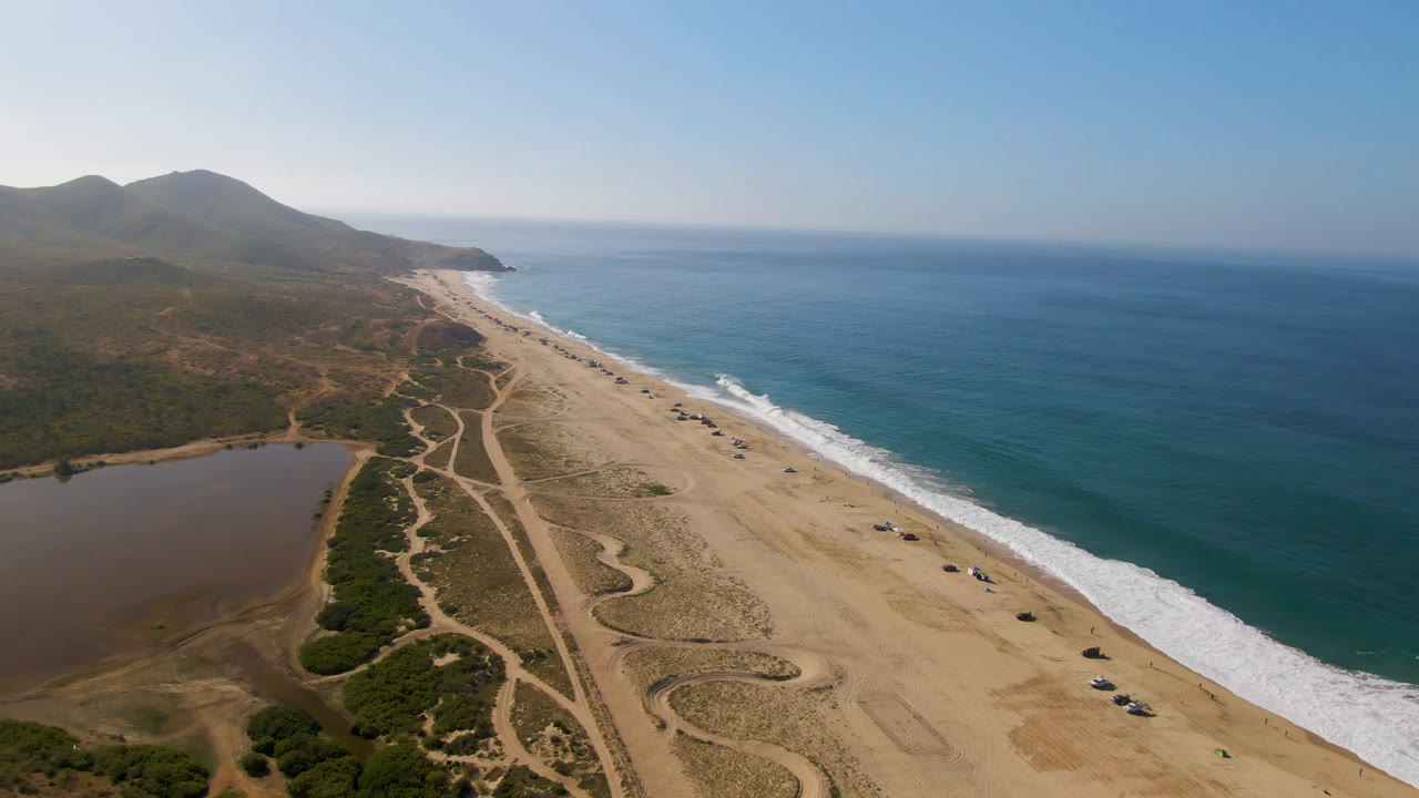 At 0:01, the aerial view in Migrino keeps the ocean to the right and the lagoon to the lower left, with pale sand, winding tracks, distant hills, and no visible people.