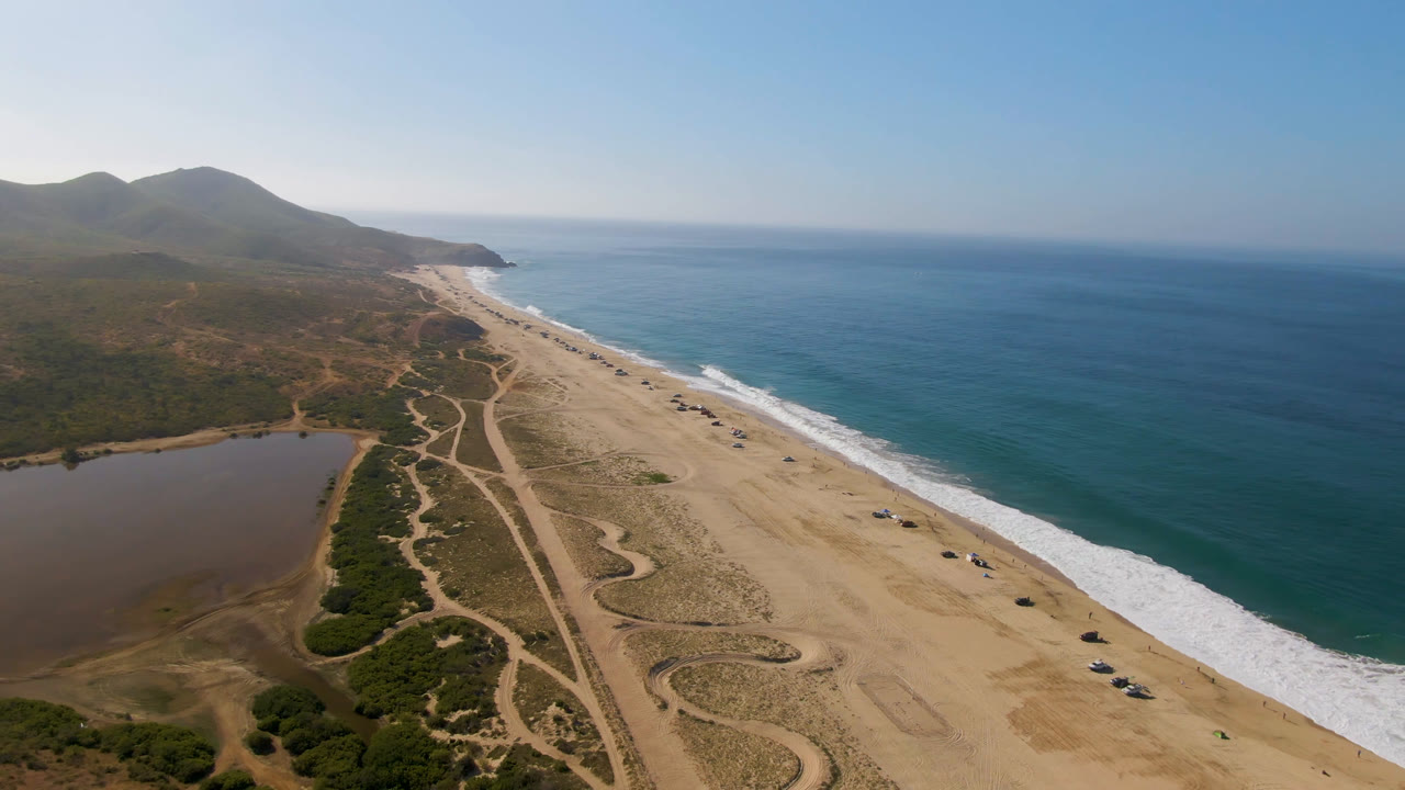 Wide aerial coastline with beach, surf line, lagoon edge, scrub, and hills.