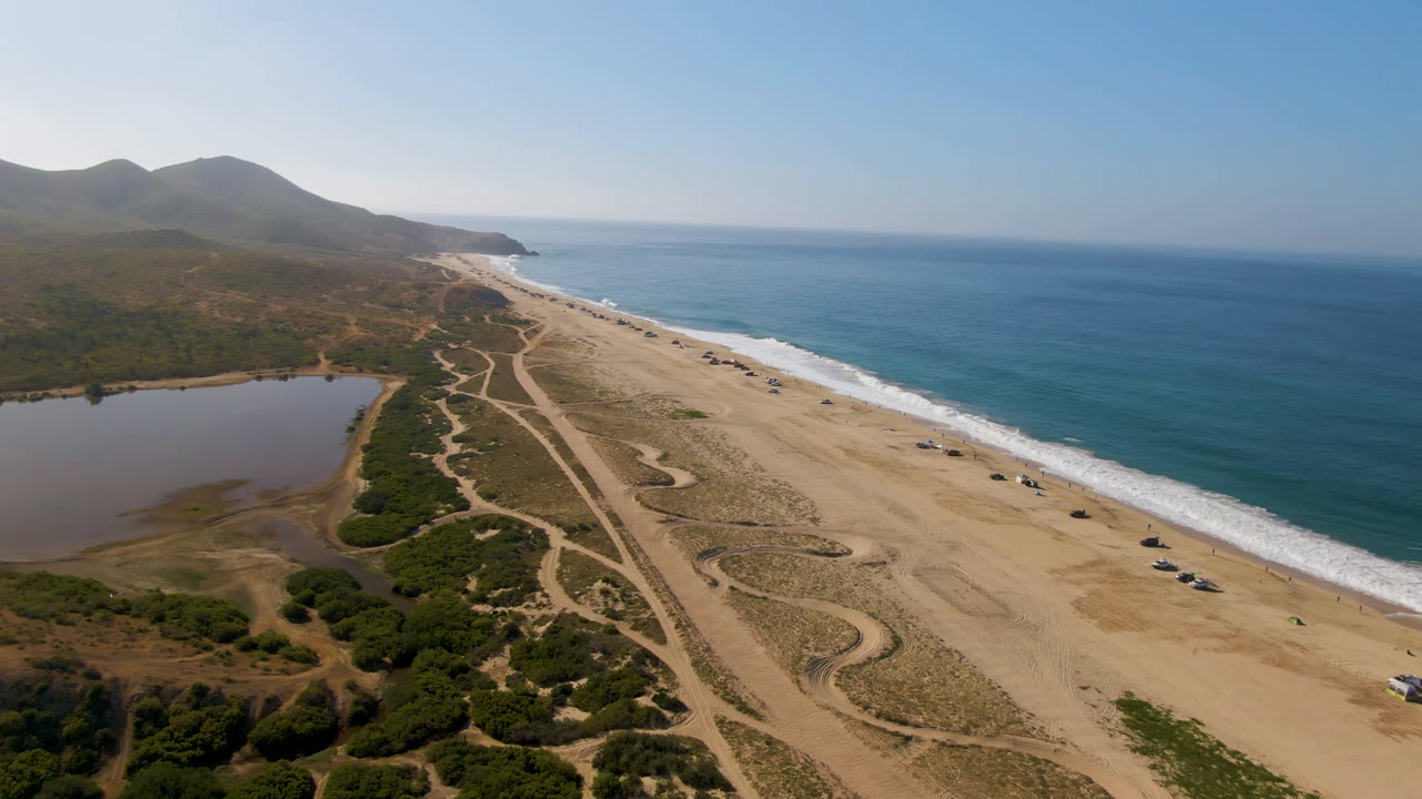 Aerial frame over Migrino, Baja California Sur, showing a sandy beach beside blue ocean, an inland body of water at left, dirt tracks, scrub, distant hills, and no visible people.