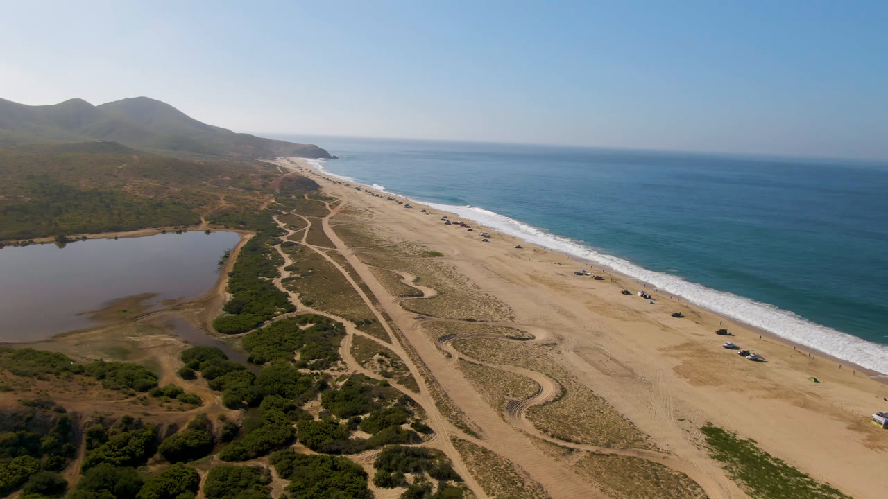 Drone-height view in Migrino, Baja California Sur, with the shoreline running diagonally, white surf on the right, a still inland water area on the left, dirt routes through scrub, and no visible people.