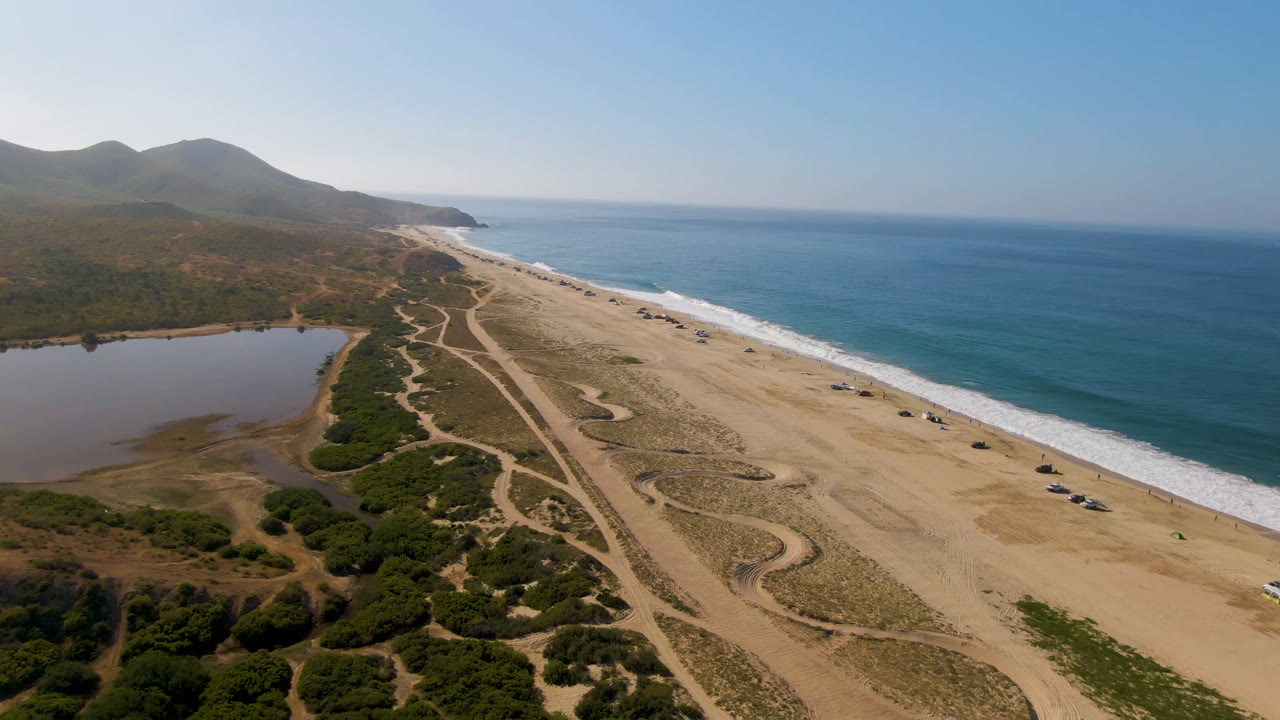 Wide drone view of ocean shoreline, beach, scrub, dirt paths, inland water, and hills.