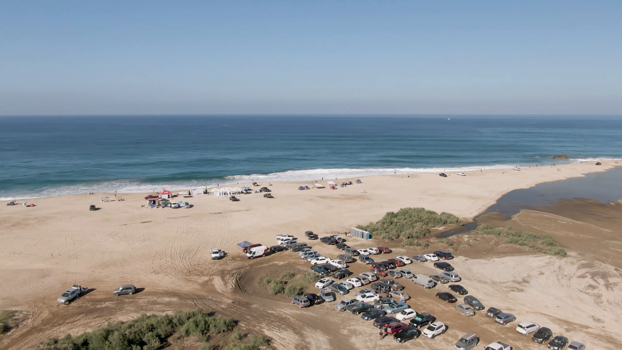 Aerial frame over Migrino, Baja California Sur, showing parked cars clustered on beach sand, low dunes in the foreground, blue ocean beyond the surf, and no visible people.