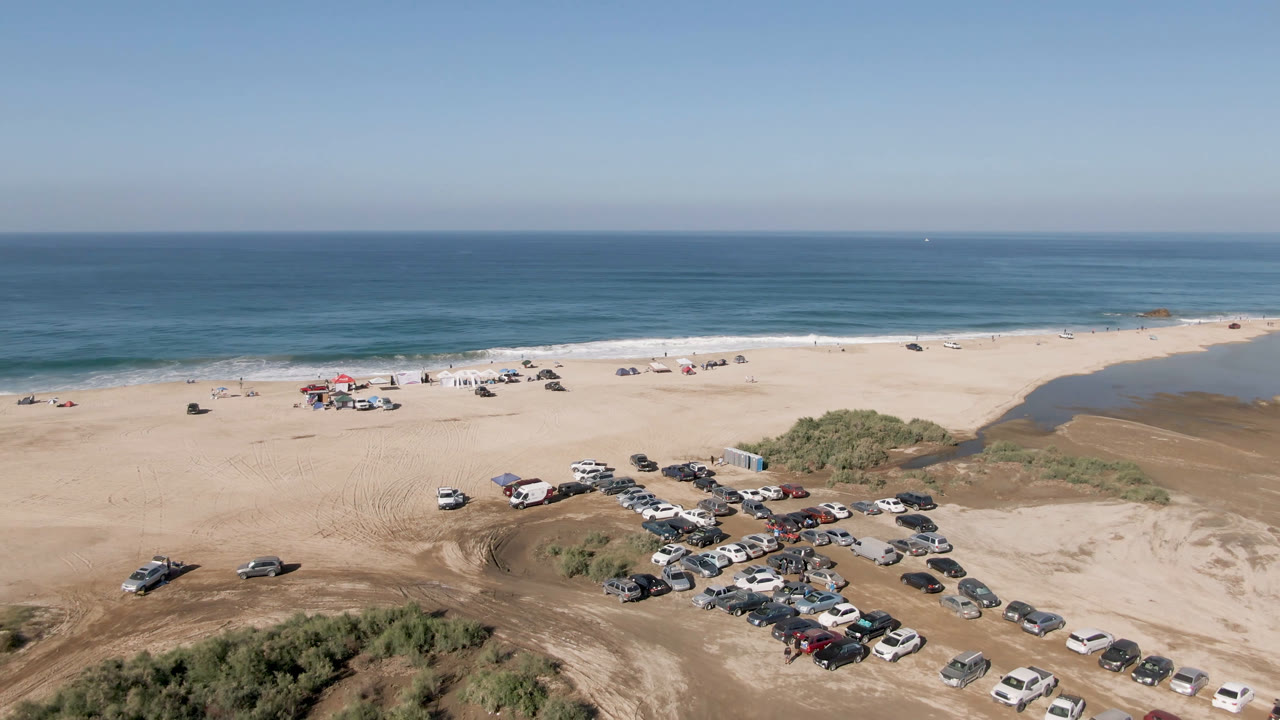 Wide aerial view in Migrino with rows of vehicles on the sand, a scrub-covered dune mound in the lower frame, a pale beach, blue water, and no visible people.
