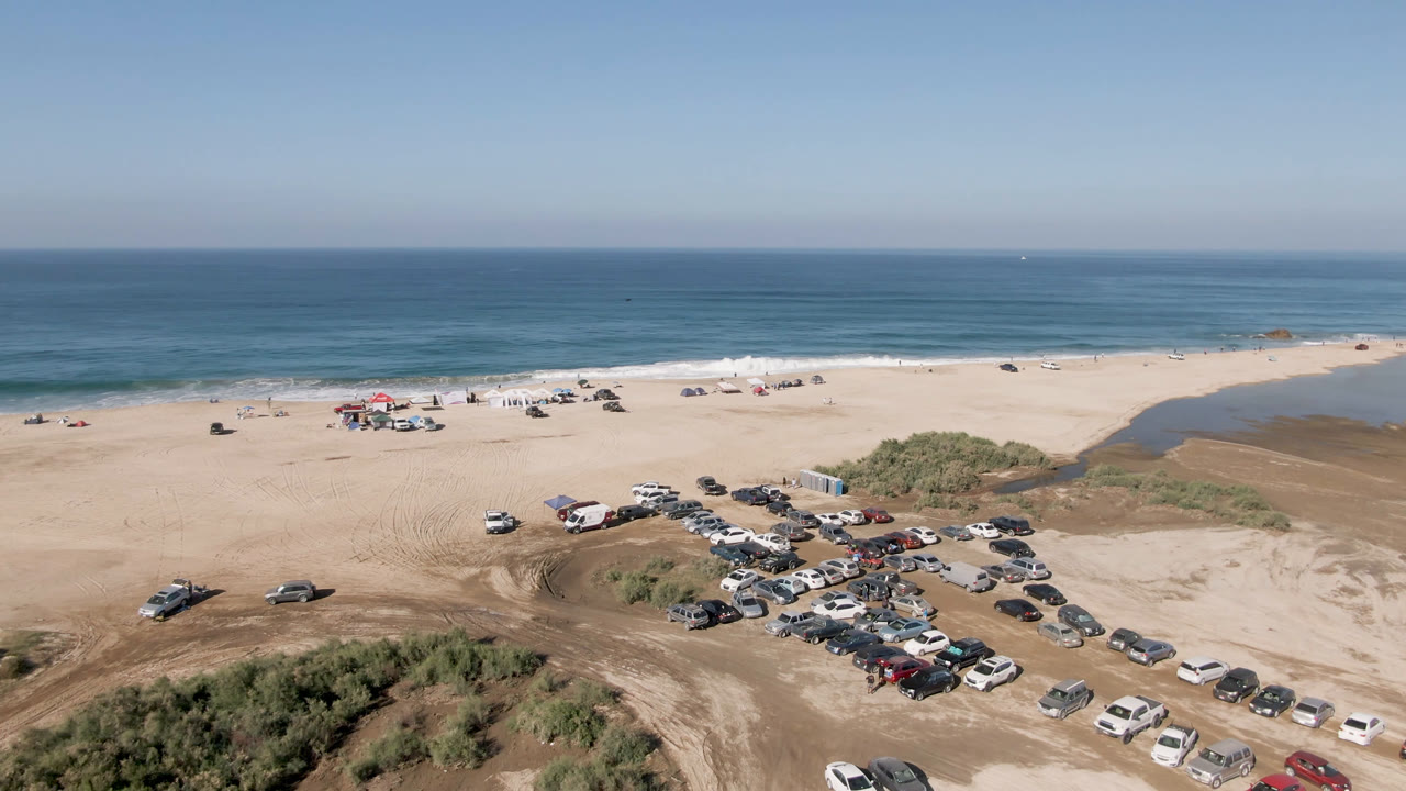 Aerial beach scene with cars, sand tracks, dunes, and ocean horizon.