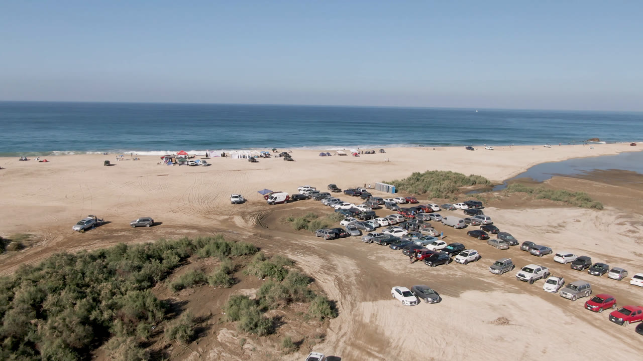 Opening aerial frame over Migrino, Baja California Sur, with rows of cars on sandy beach access, dune brush in front, blue ocean beyond, and no visible people.