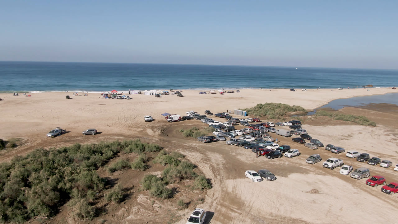 Aerial frame in Migrino showing clustered vehicles on pale sand, a dark water channel near the shoreline, low dunes in the foreground, and no visible people.